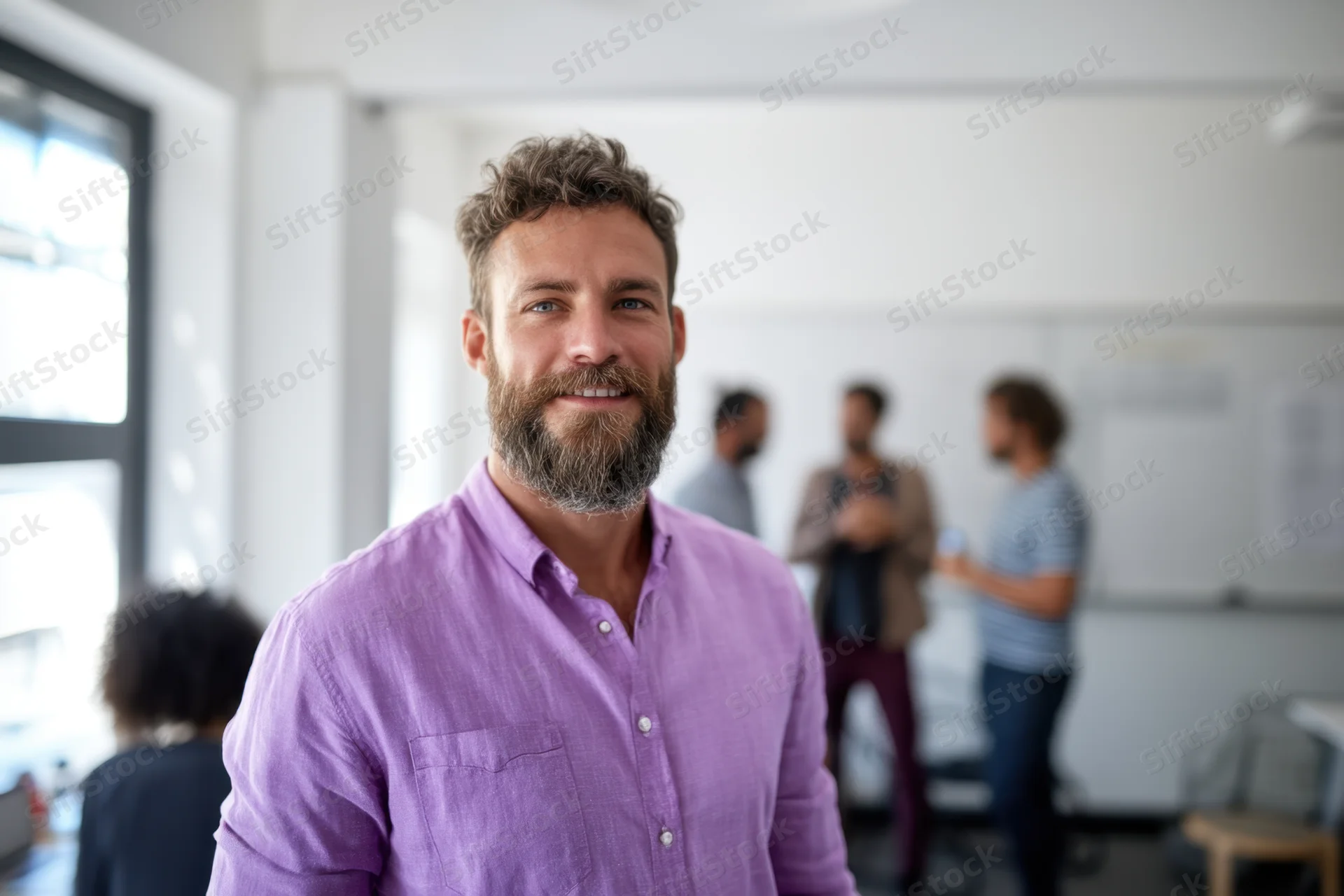 Smiling bearded man in purple shirt standing in office with colleagues in background