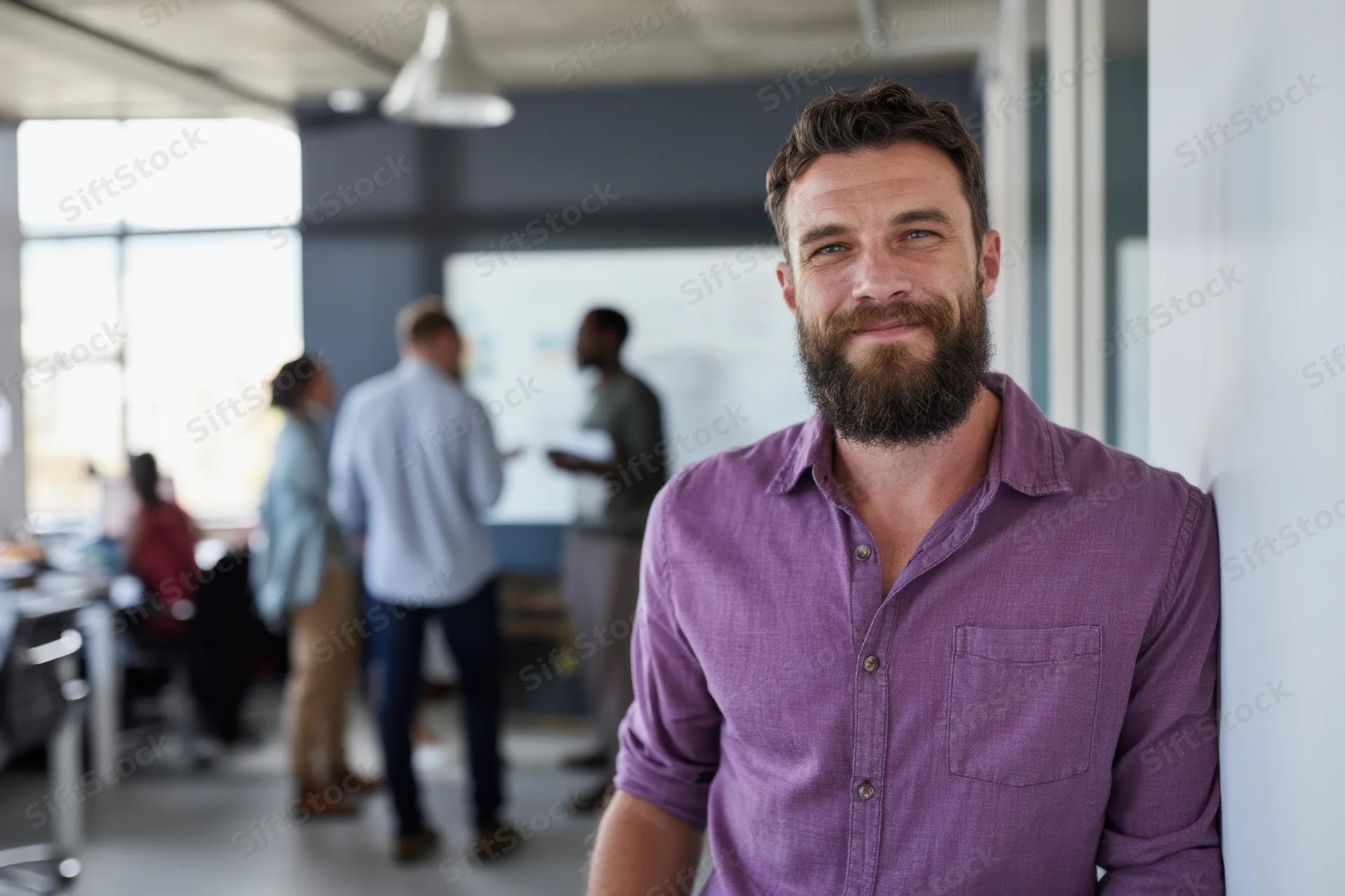 Bearded man in purple shirt smiling while leaning against wall in modern office with colleagues collaborating in background