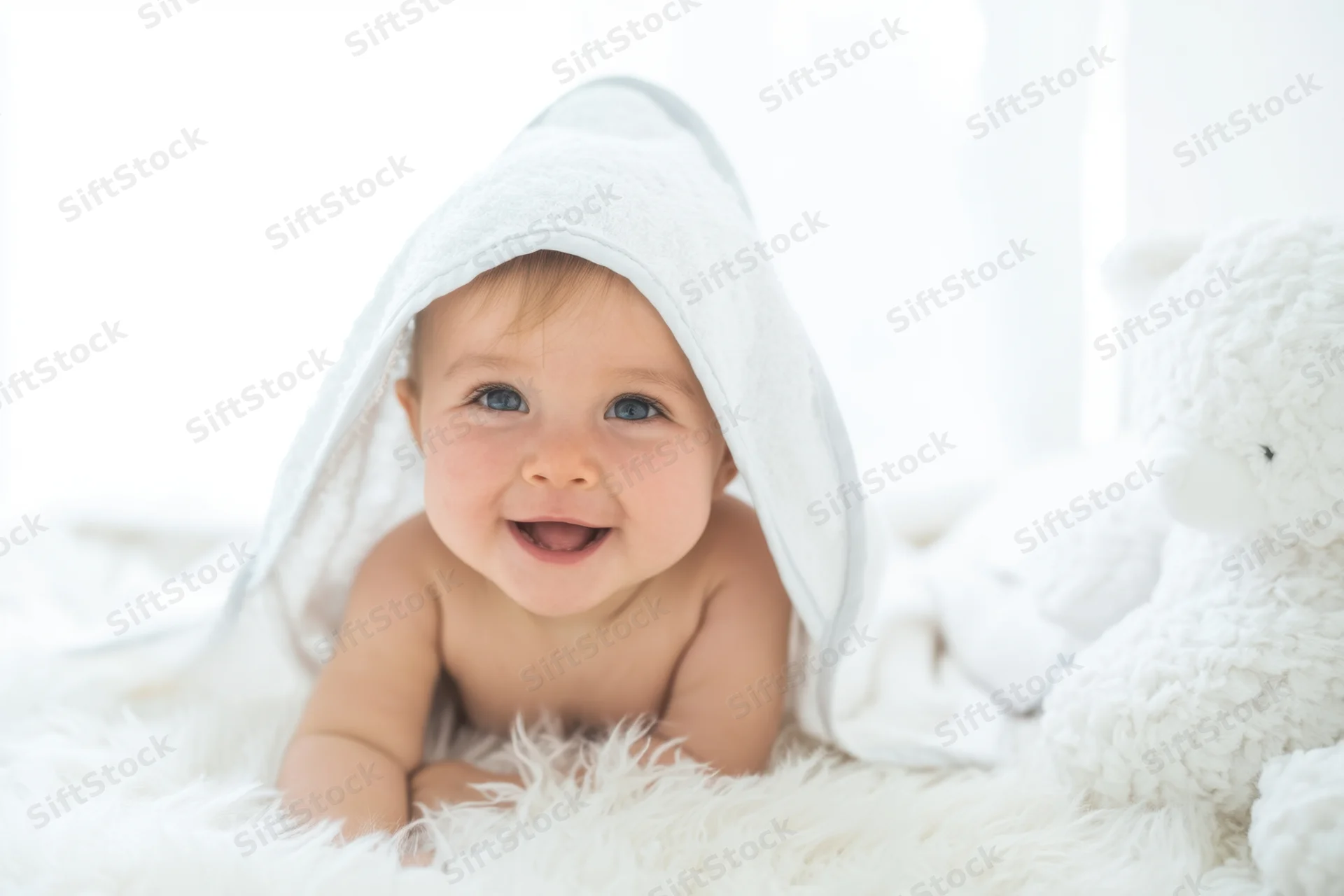 Happy baby smiling with white hooded towel over head lying on fluffy white rug beside plush teddy bear