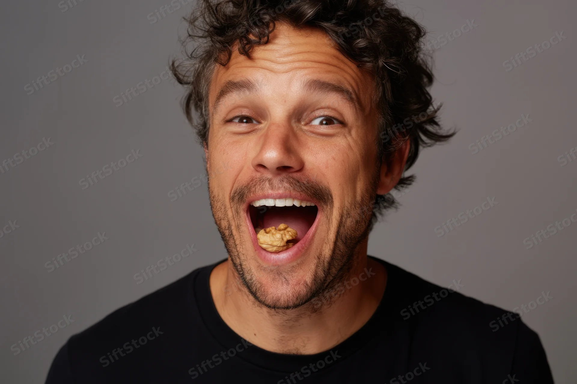 Man with curly hair and stubble smiling widely with walnut in his open mouth against gray background