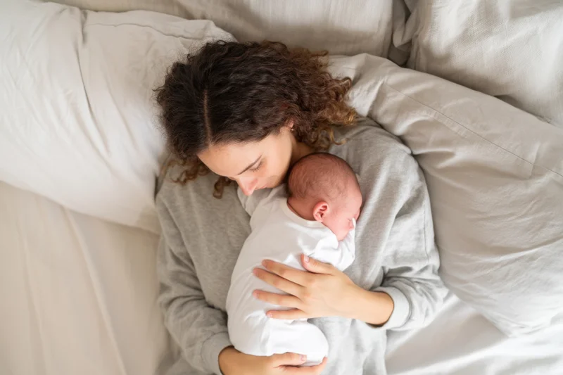 Mother cradling sleeping newborn in bed, wrapped in white swaddle, wearing gray loungewear, surrounded by soft bedding