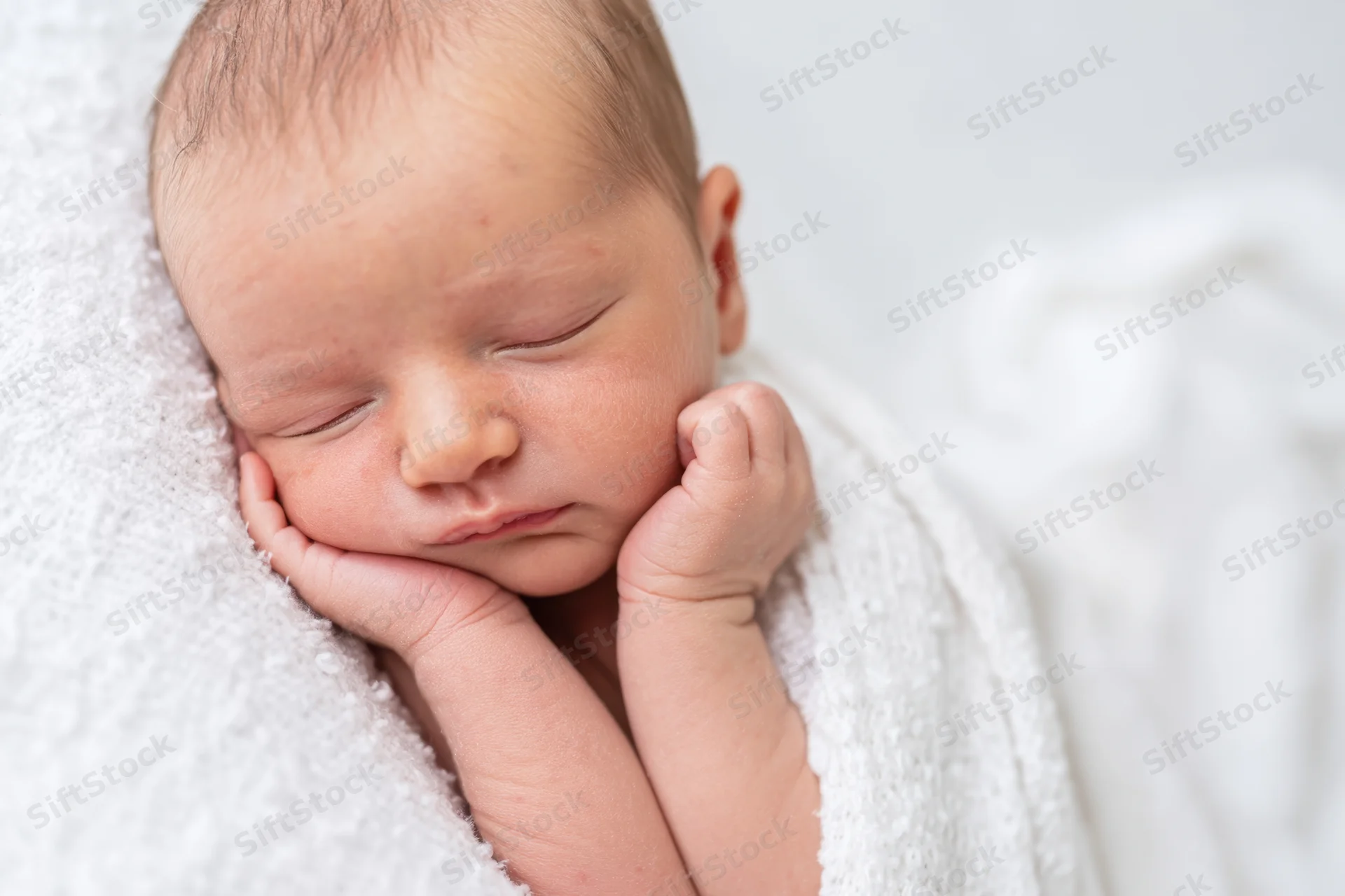 Newborn baby sleeping with hands near cheeks wrapped in white blanket