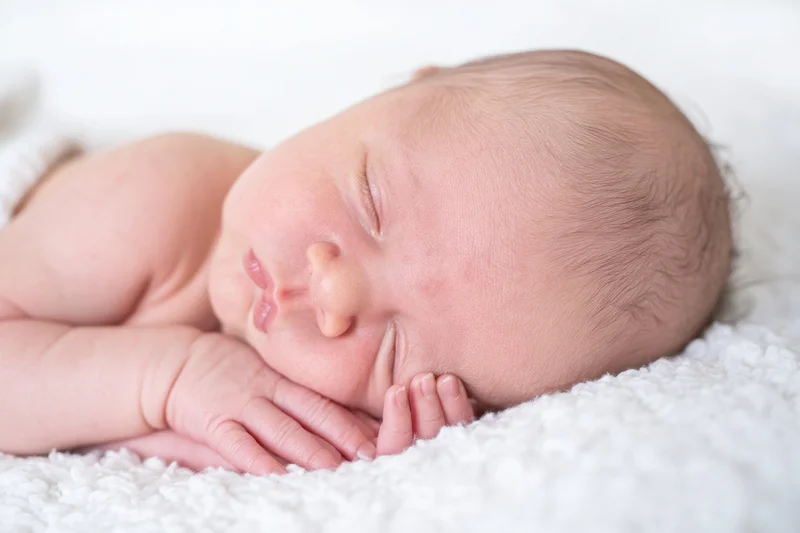 Newborn baby sleeping with hands folded near cheeks on white textured blanket