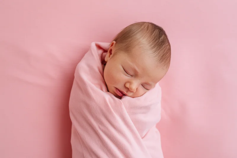 Newborn baby sleeping swaddled in pink blanket on soft pink surface