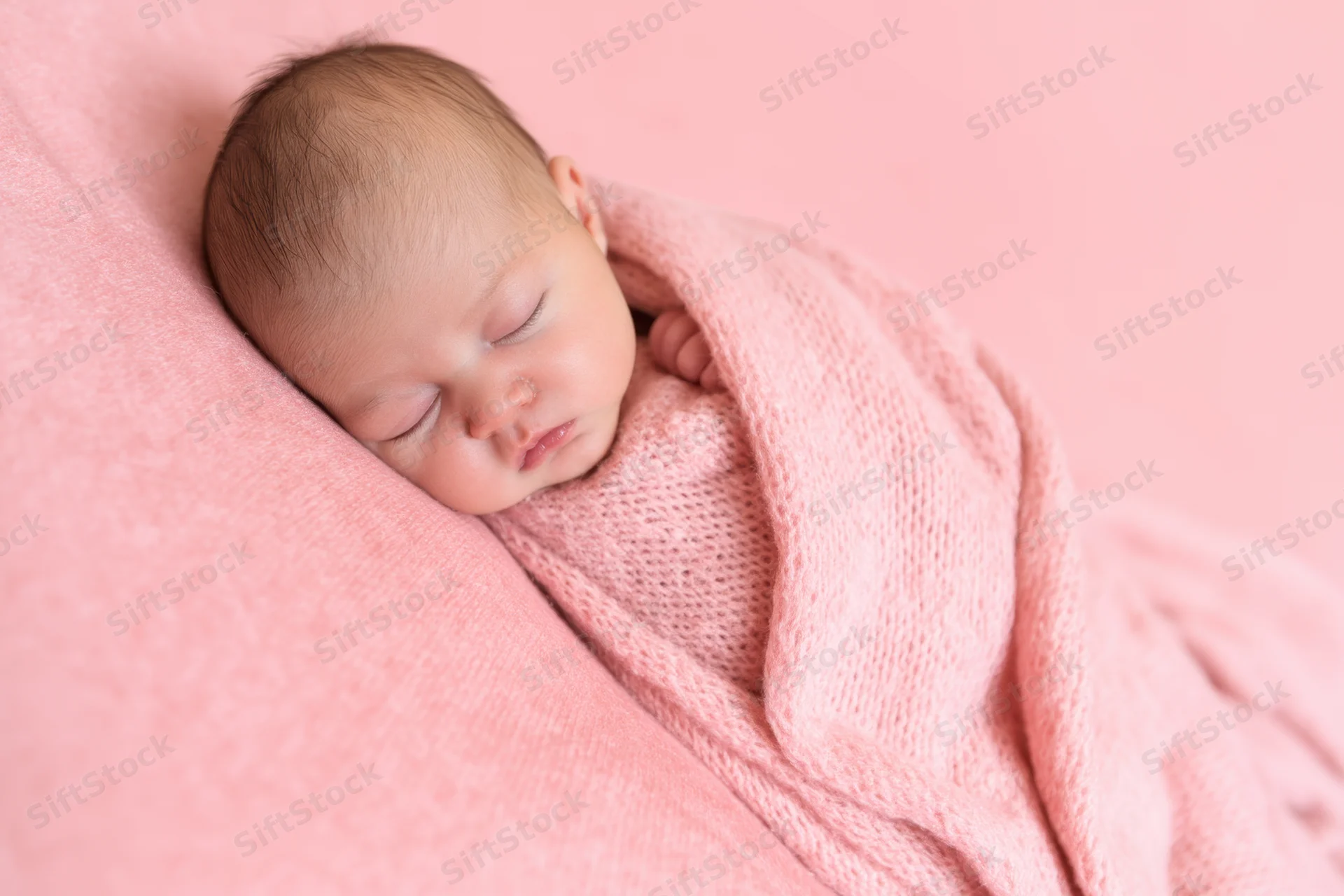 Newborn baby sleeping swaddled in soft pink blanket on pink background