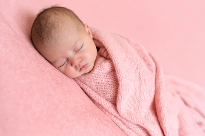 Newborn baby sleeping swaddled in soft pink blanket on pink background
