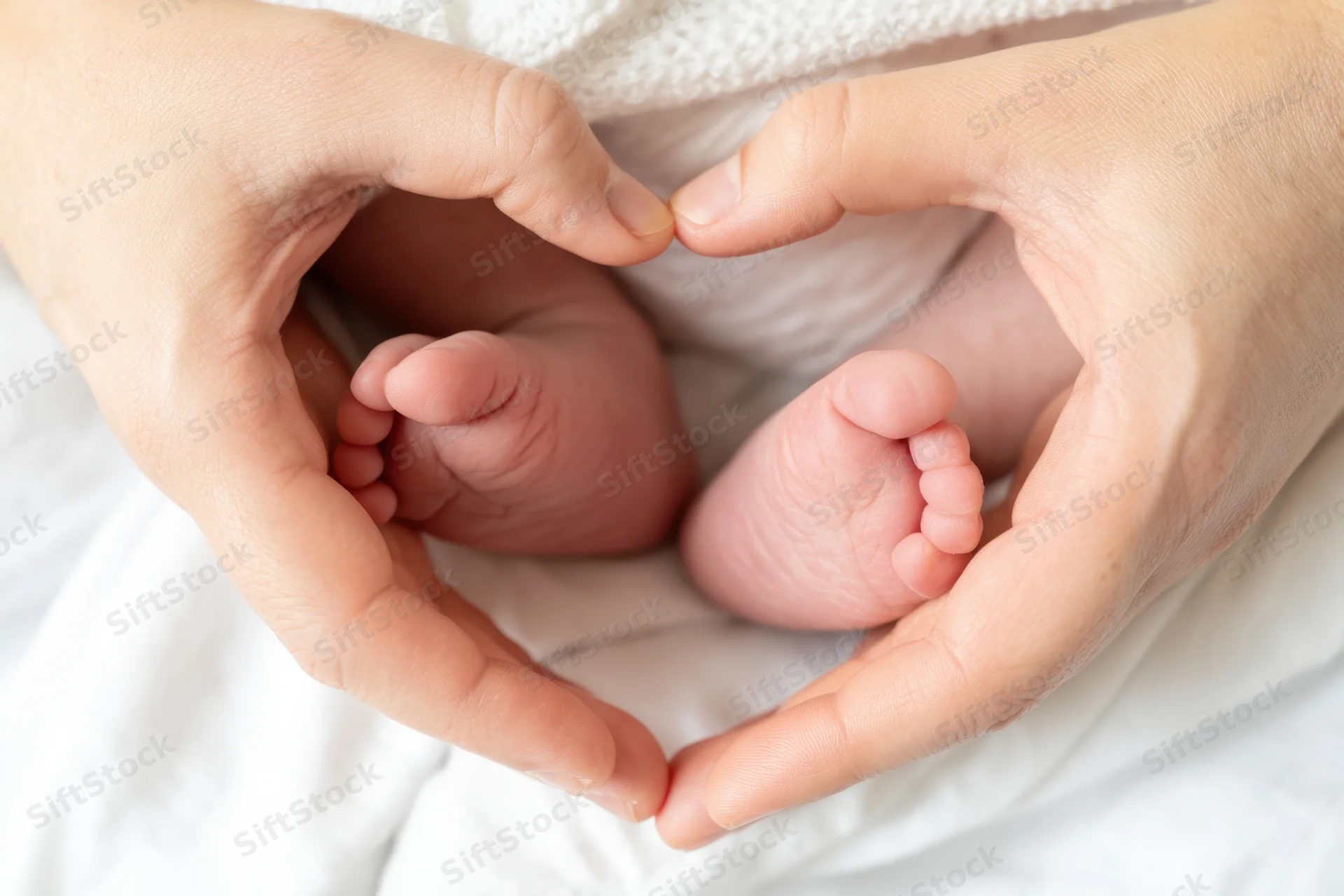 Adult hands forming a heart shape around a newborn baby's feet on a white blanket