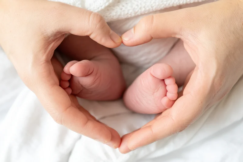 Adult hands forming a heart shape around a newborn baby's feet on a white blanket