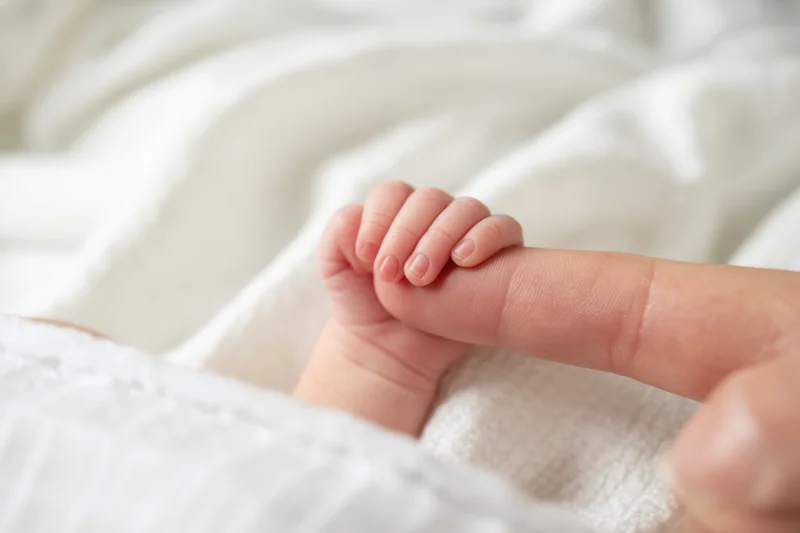 Newborn baby's tiny hand gripping an adult finger on a white blanket