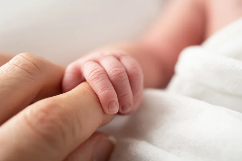 Newborn baby's tiny hand gripping an adult finger resting on a white blanket