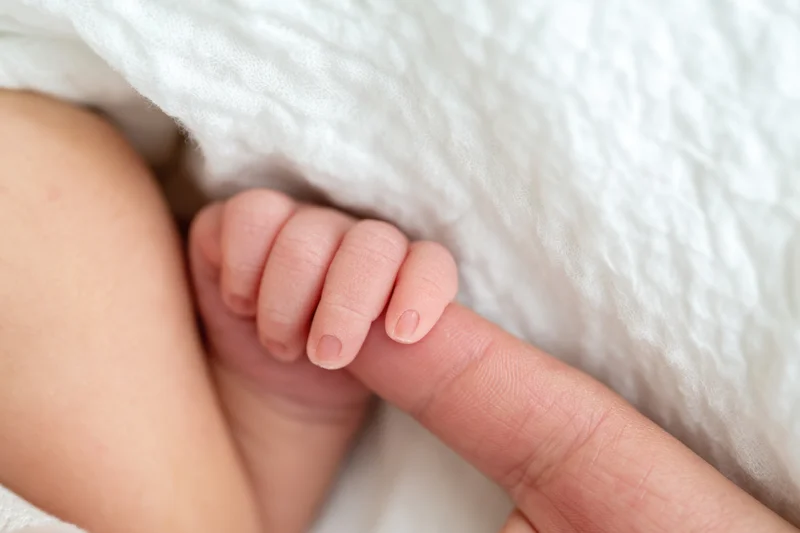 Newborn baby's tiny hand gripping an adult finger resting on a white blanket