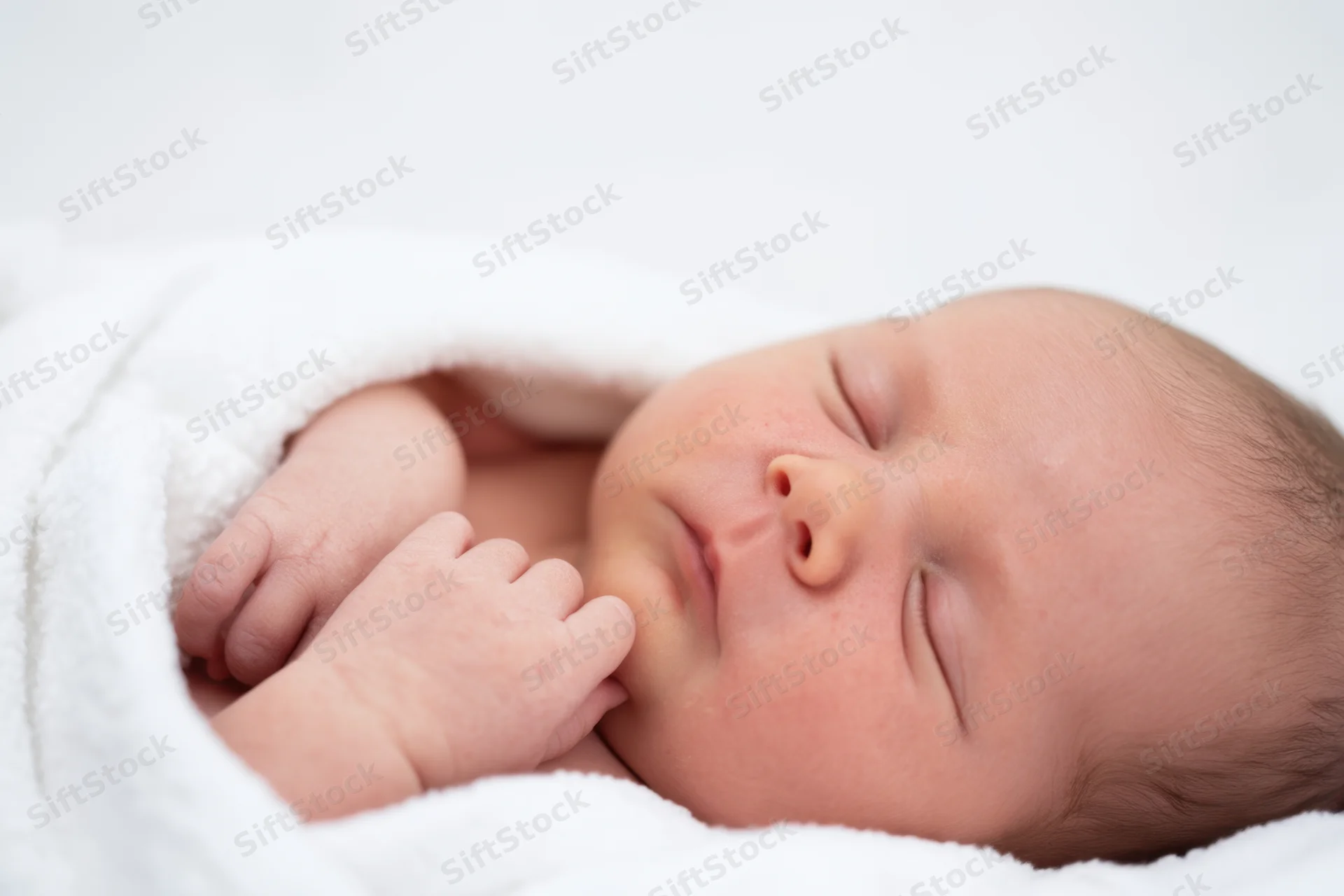 Newborn baby sleeping peacefully wrapped in white blanket with rosy cheeks and tiny fingers near face