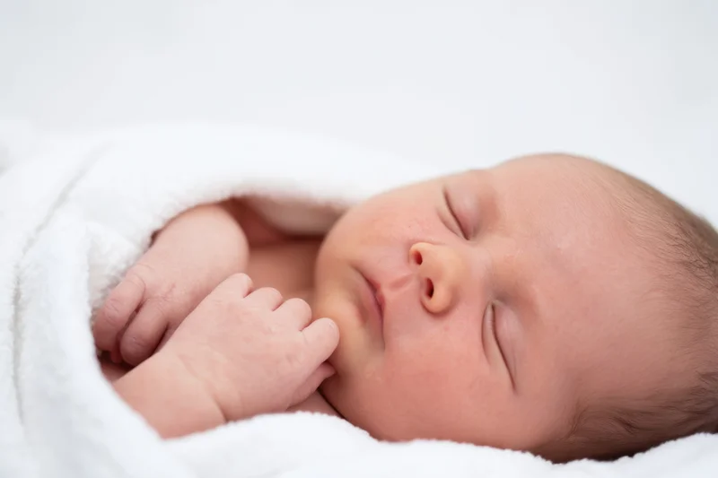 Newborn baby sleeping peacefully wrapped in white blanket with rosy cheeks and tiny fingers near face