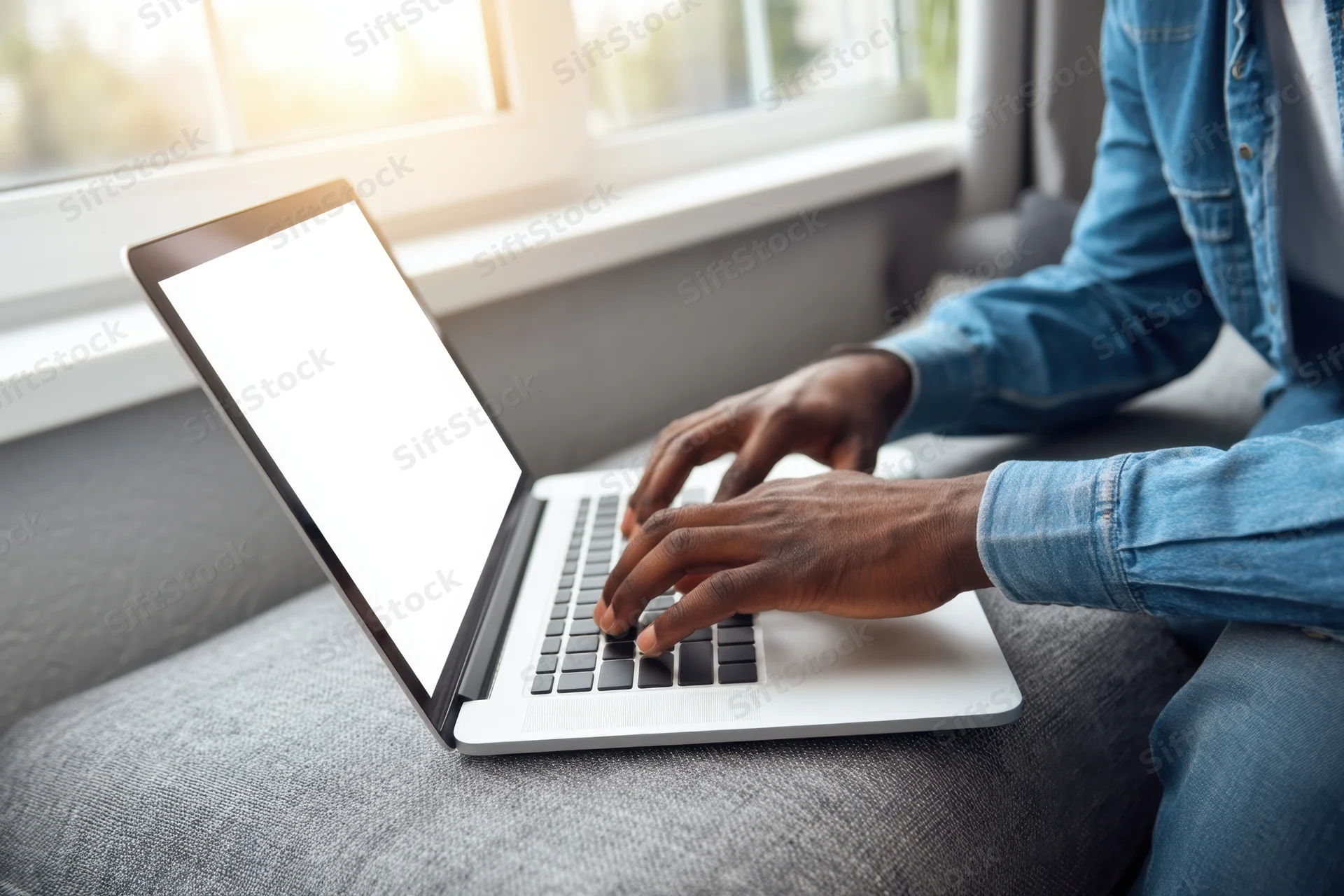 Man typing on laptop while sitting on gray couch near sunlit window