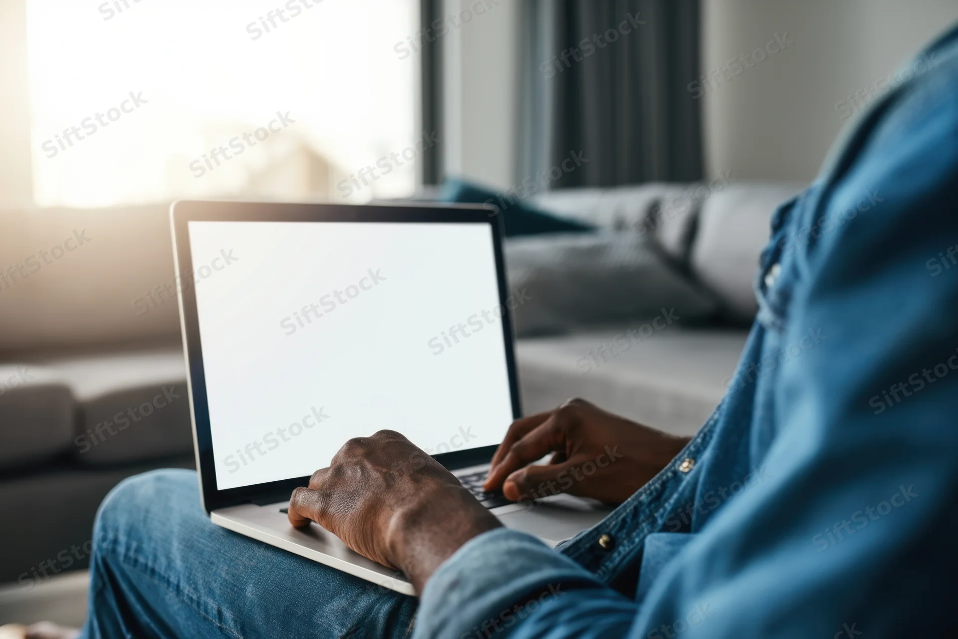 Man typing on laptop with blank screen while sitting on gray couch near sunlit window