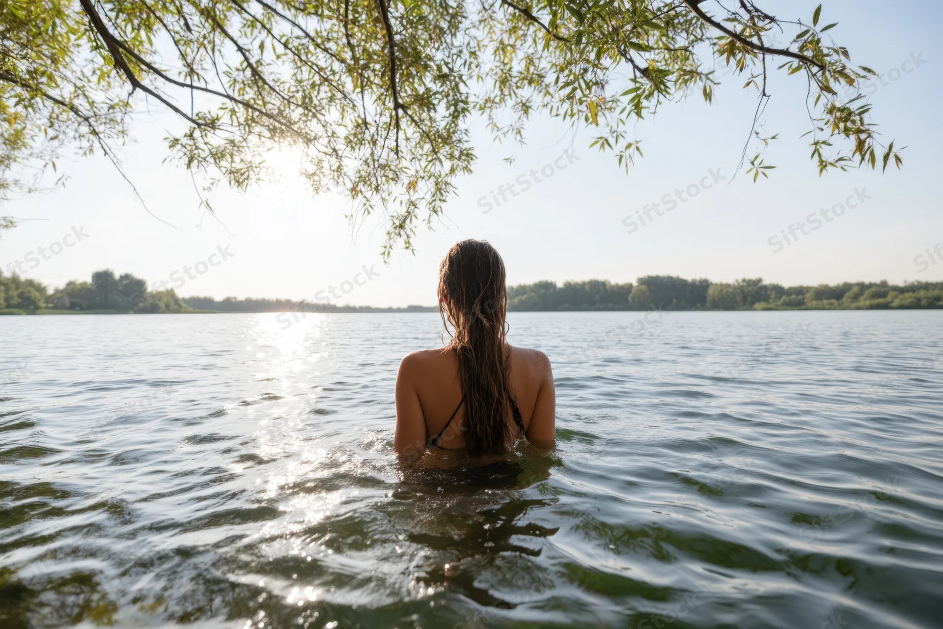 Woman standing chest-deep in a tranquil lake with wet hair, back to camera, under overhanging tree branches, sunlight glinting on water surface