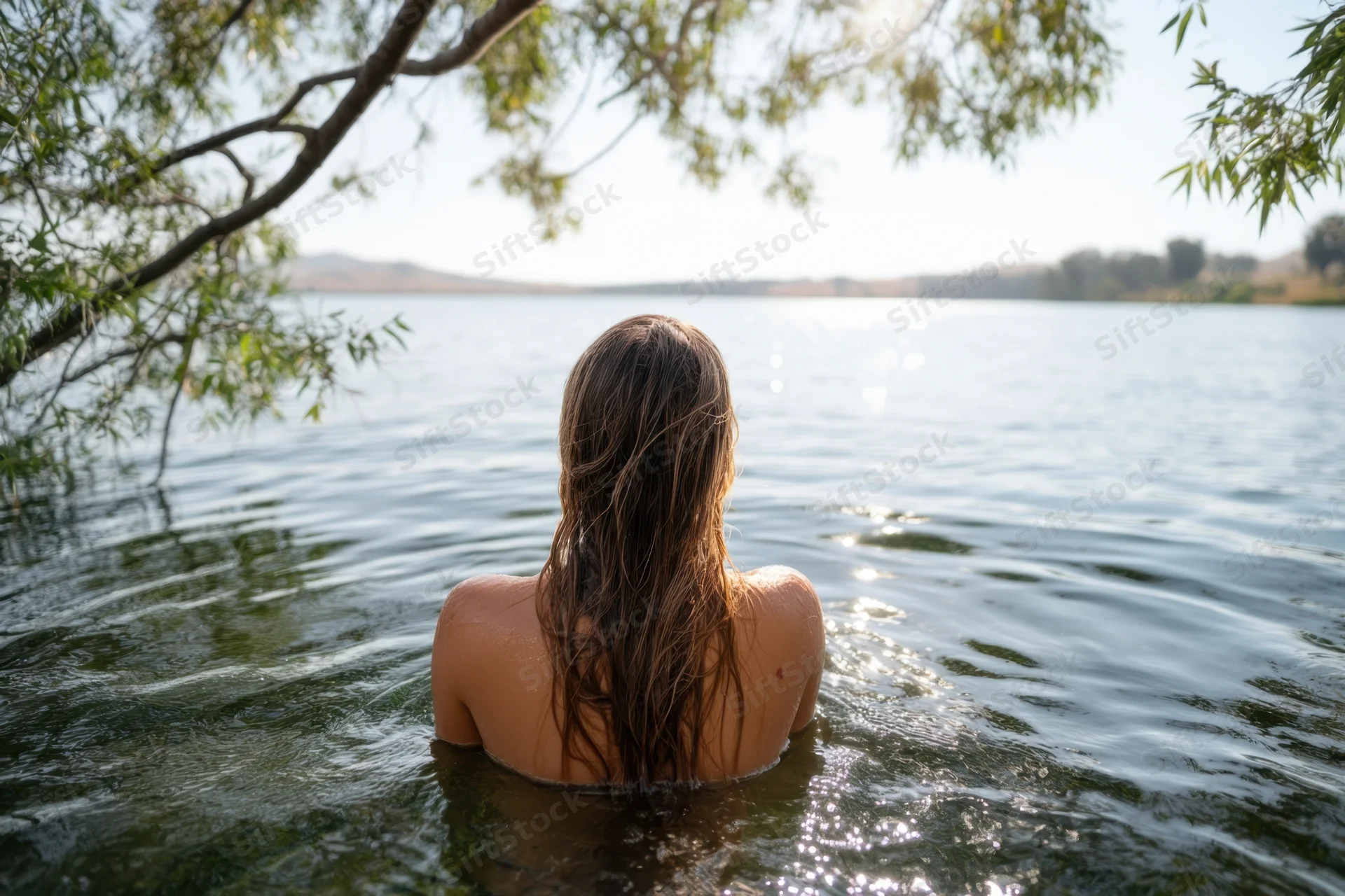 Woman standing chest-deep in a tranquil lake with wet hair, back to camera, surrounded by trees and sunlight