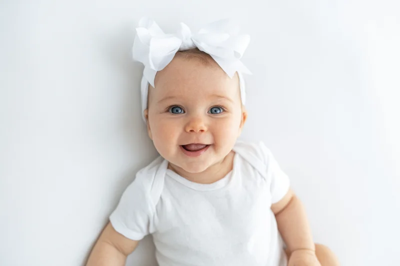 Smiling baby in white onesie with large white bow on headband, studio portrait
