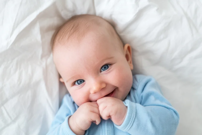 Smiling baby lying on white bed with hands near mouth wearing light blue onesie