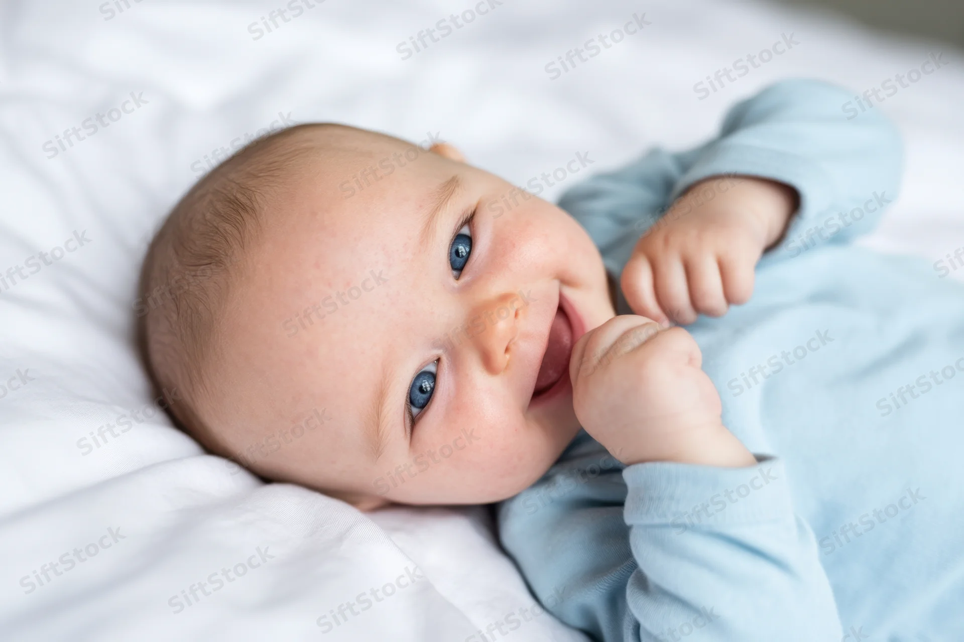 Smiling baby lying on white bed wearing light blue onesie