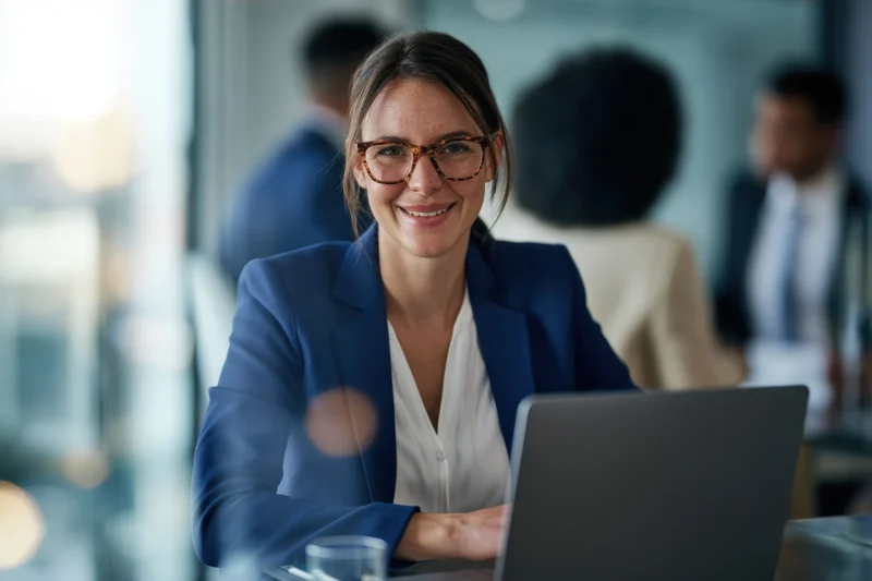 Woman in blue blazer smiling at laptop in modern office setting
