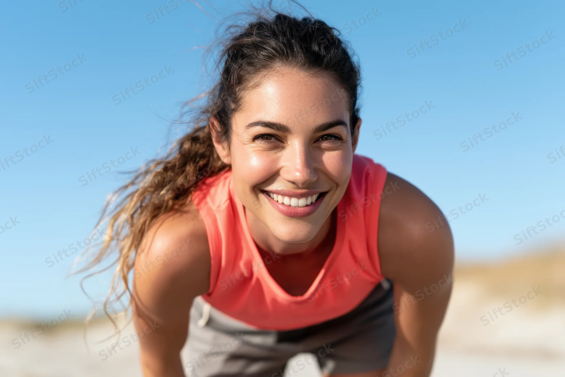 Smiling woman in coral athletic wear leaning forward on a sandy beach under clear blue sky