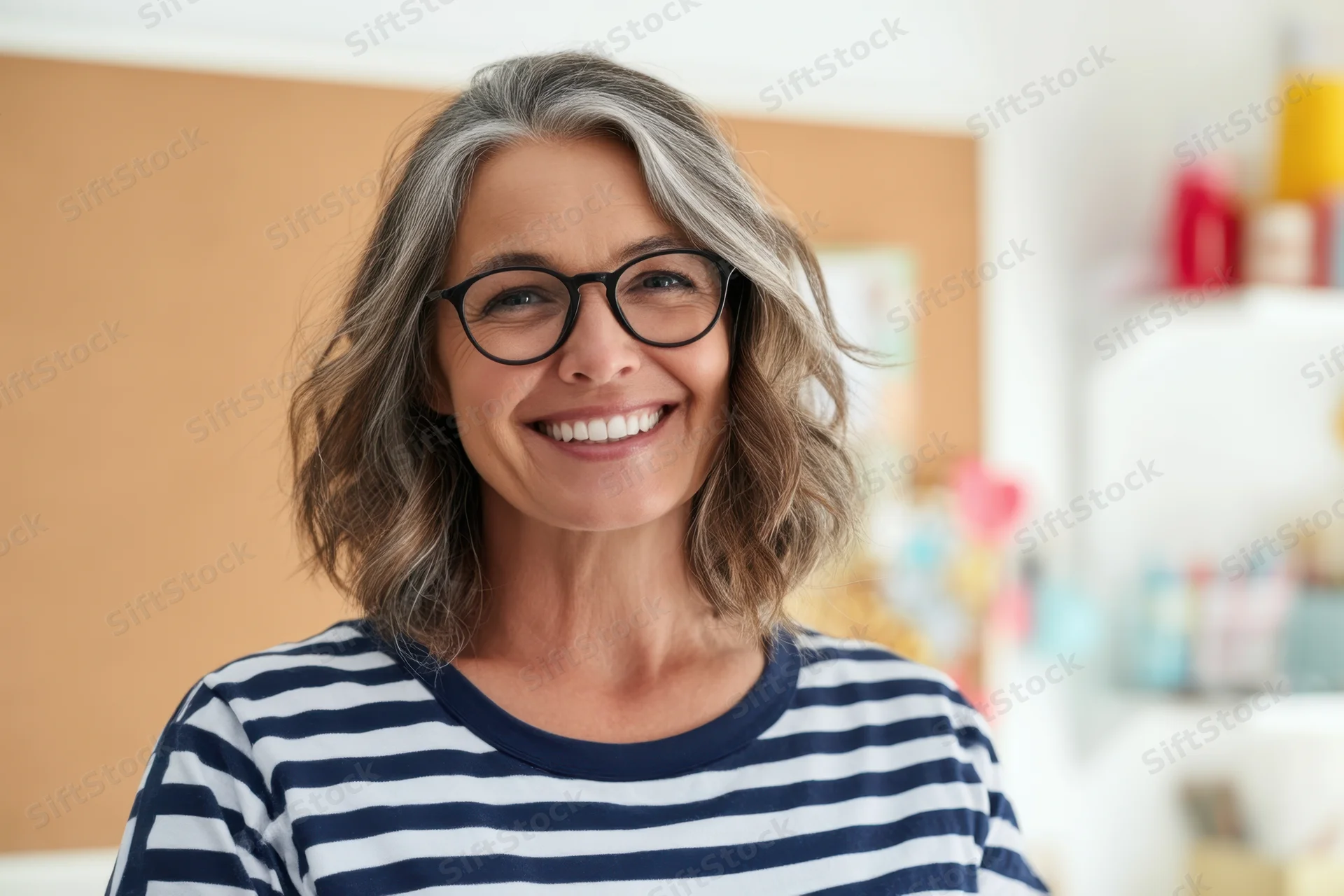 Smiling middleaged woman with gray hair and black-rimmed glasses wearing striped shirt in classroom setting