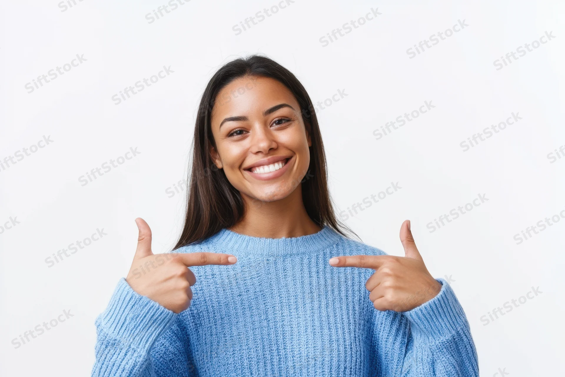 Young woman smiling and pointing at herself with thumbs up in a blue sweater against white background
