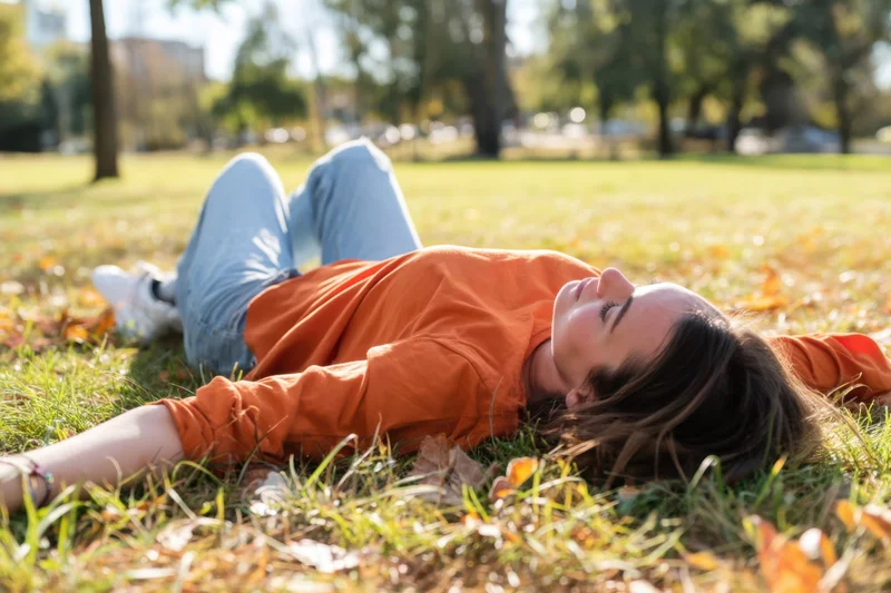 Woman lying on sunlit grass in a park with autumn leaves around her