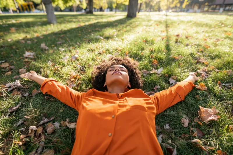 Woman lying on sunlit grass in a park with arms spread, surrounded by autumn leaves