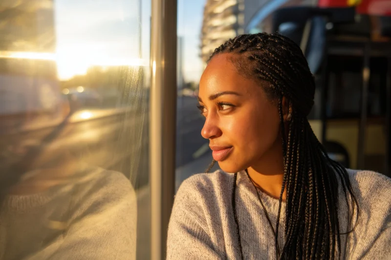 Woman with braided hair looking out a bus window during golden hour