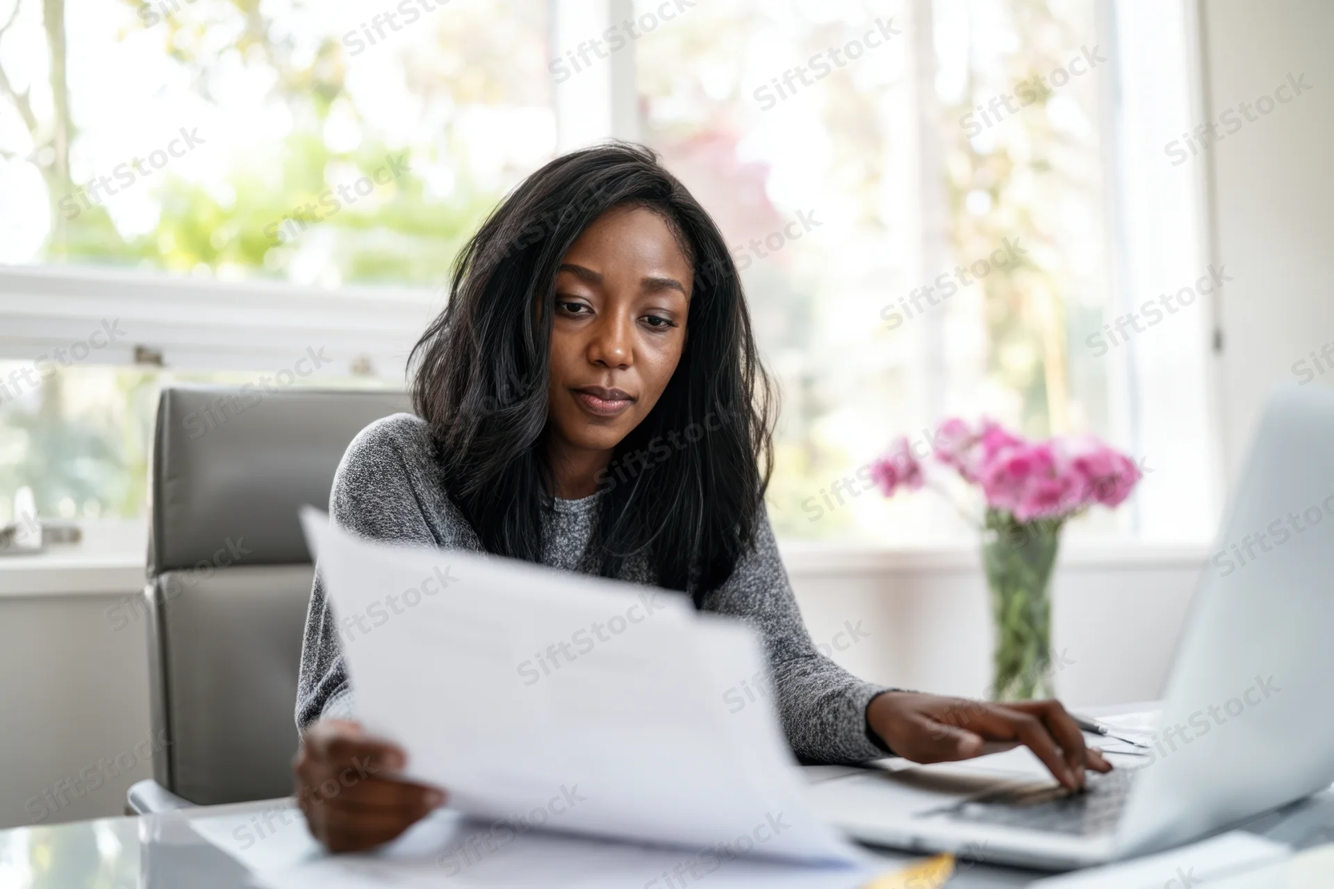 Woman reviewing document while typing on laptop at desk near window