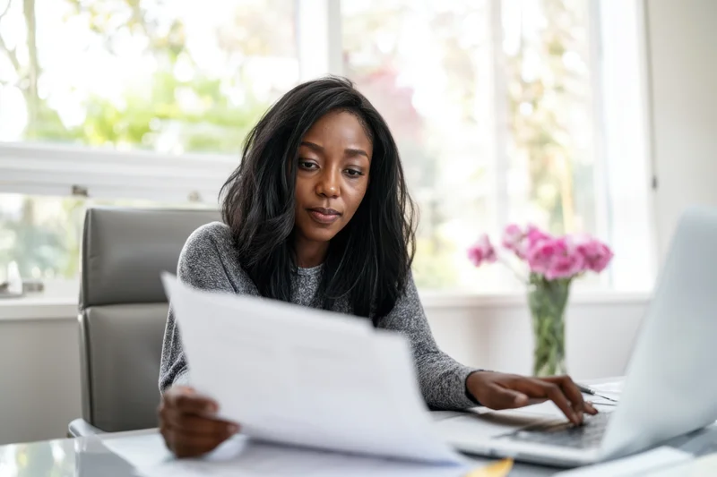Woman reviewing document while typing on laptop at desk near window