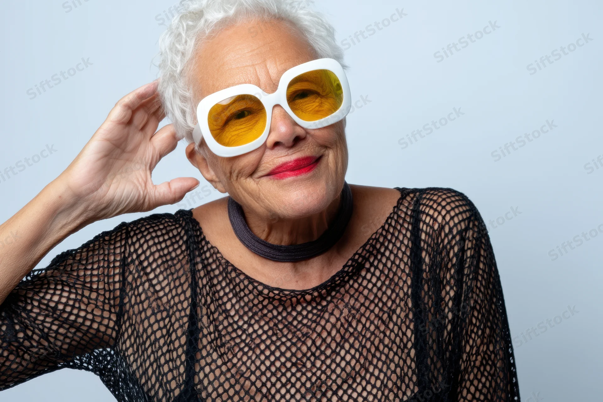 Senior woman with short white hair wearing oversized white-framed yellow sunglasses, smiling while touching her hair, in a studio portrait with a light blue background