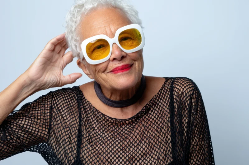 Senior woman with short white hair wearing oversized white-framed yellow sunglasses, smiling while touching her hair, in a studio portrait with a light blue background