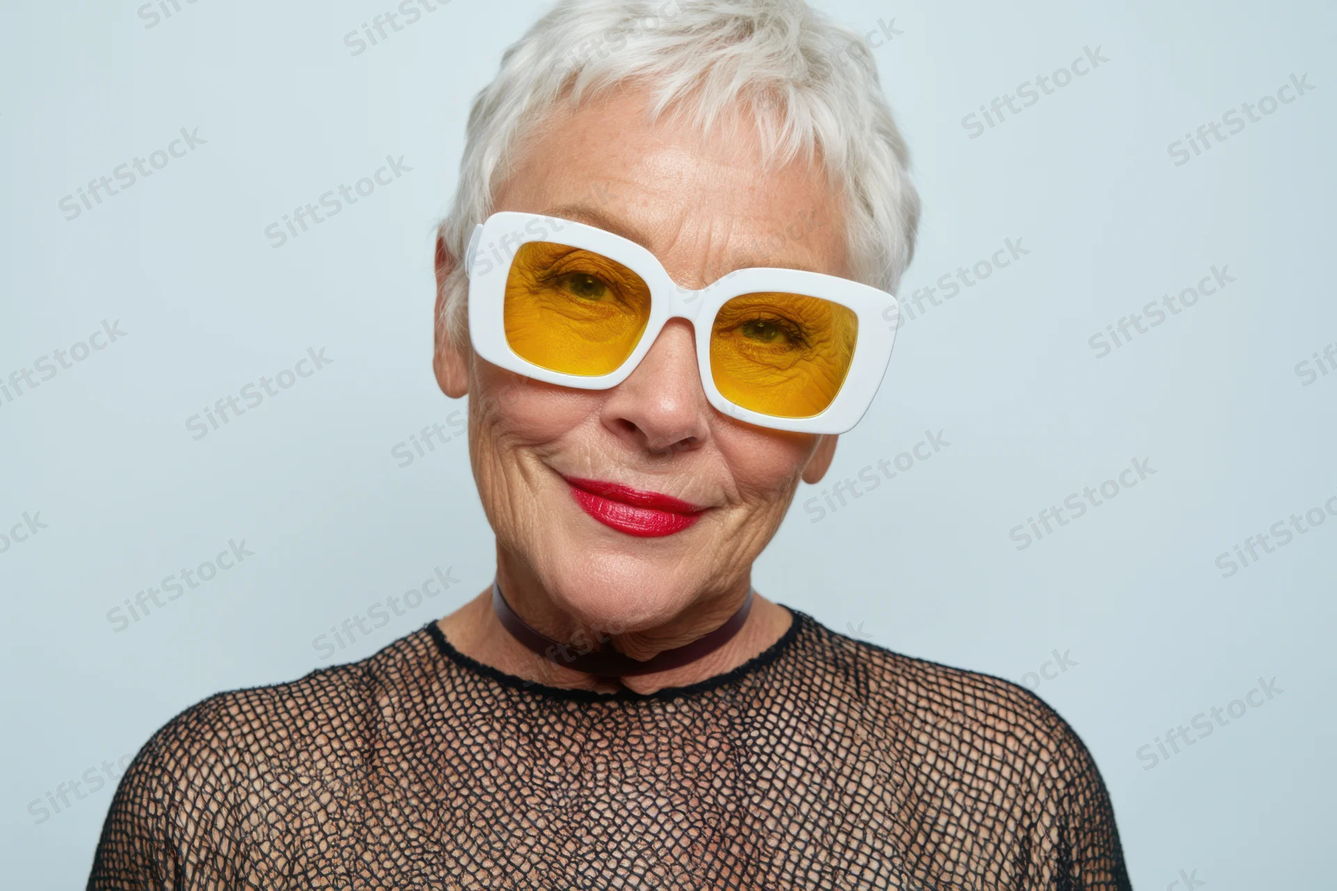 Senior woman with short white hair wearing oversized yellow-lensed sunglasses and red lipstick, smiling in a studio portrait