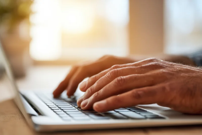 Close-up of male hands typing on a laptop keyboard at a wooden desk in a sunlit room