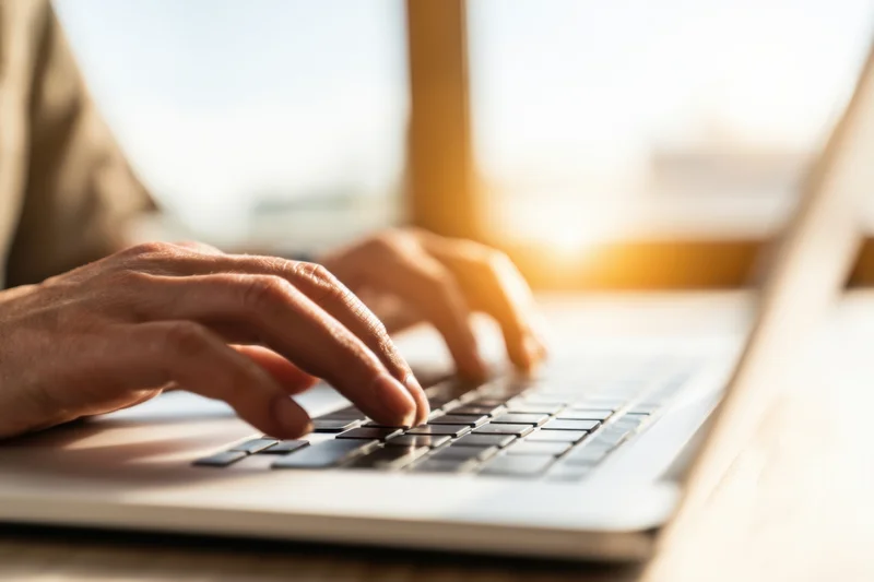Hands typing on a laptop keyboard in a sunlit indoor workspace