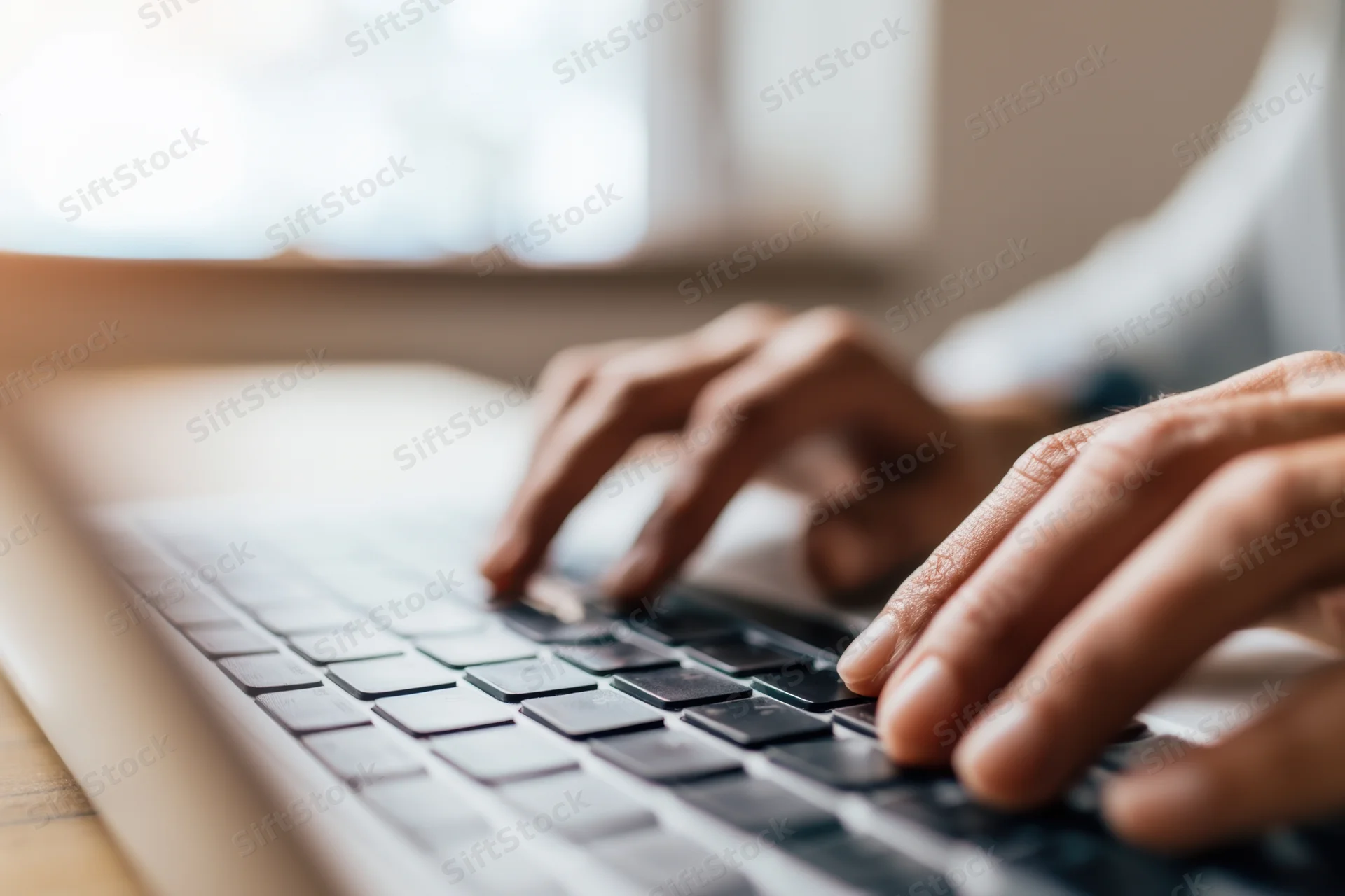 Hands typing on a laptop keyboard at a desk near a window