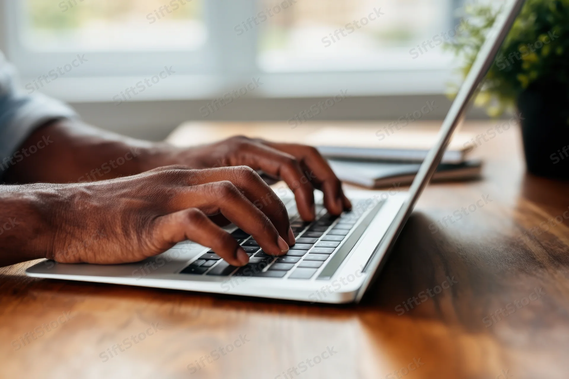 Hands typing on a silver laptop at a wooden desk by a window