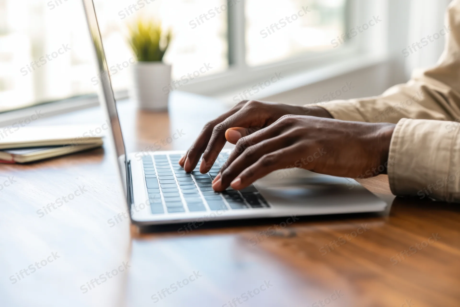 Hands typing on a silver laptop at a wooden desk by a window