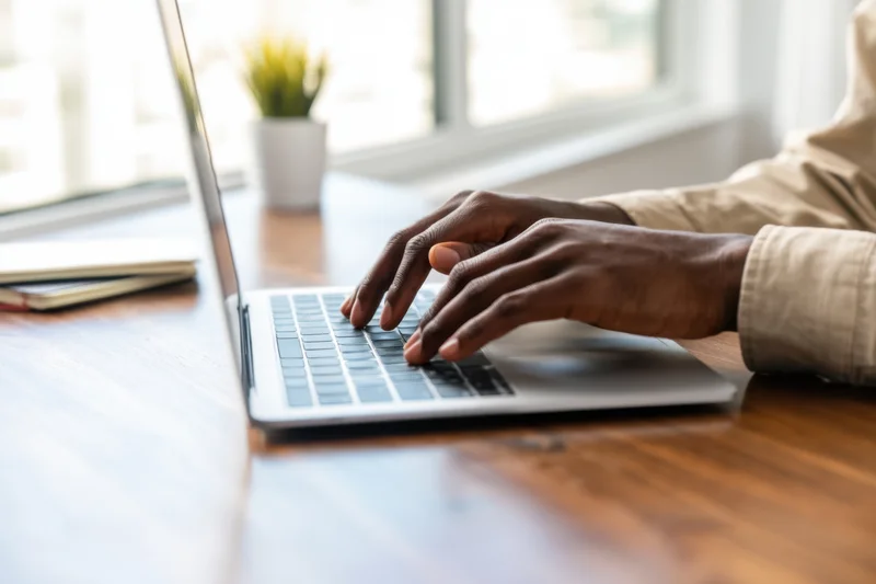 Hands typing on a silver laptop at a wooden desk by a window