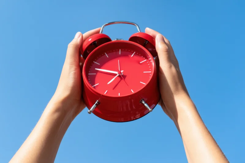 Woman's hands holding red analog alarm clock against clear blue sky
