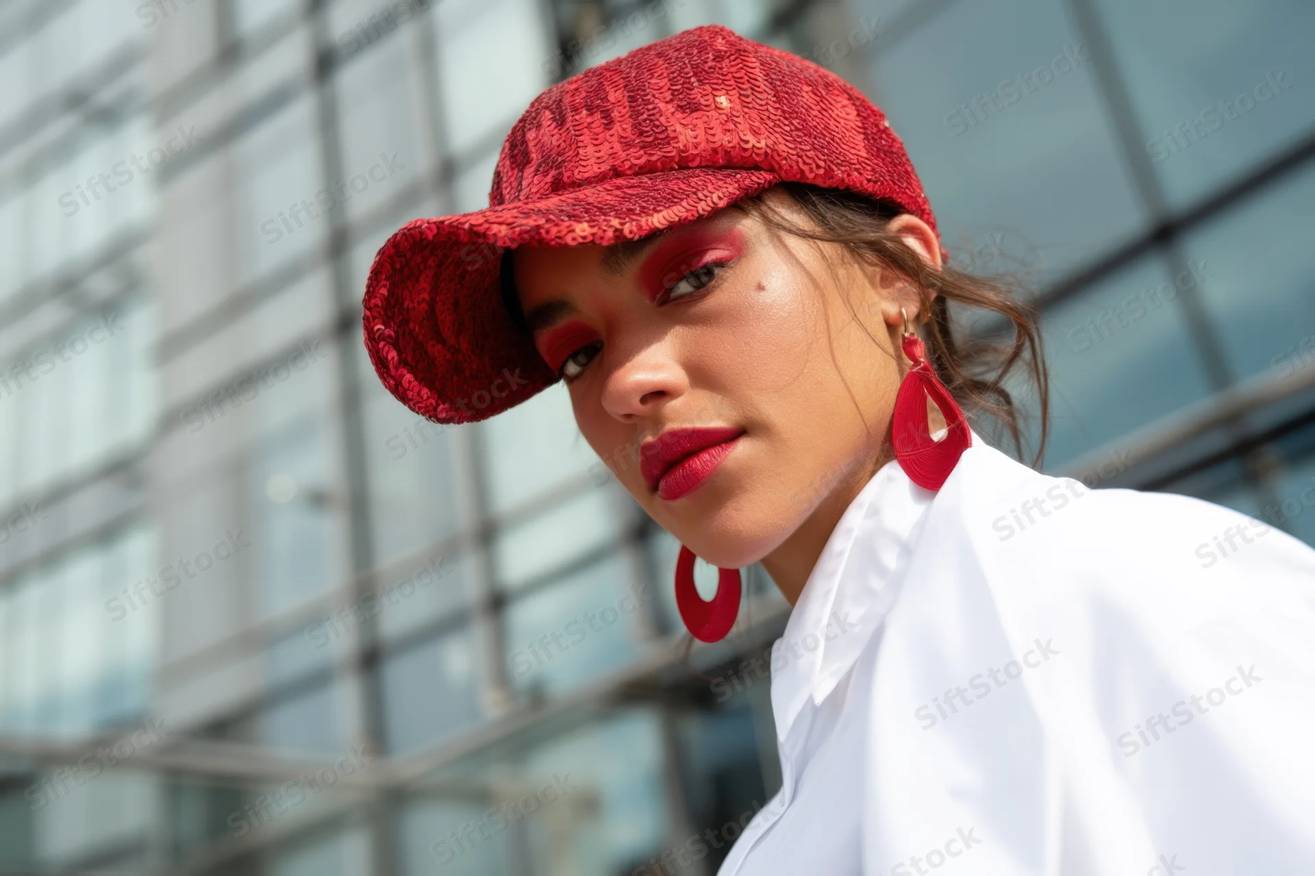 Woman in red sequined cap and bold red makeup posing outdoors near glass building