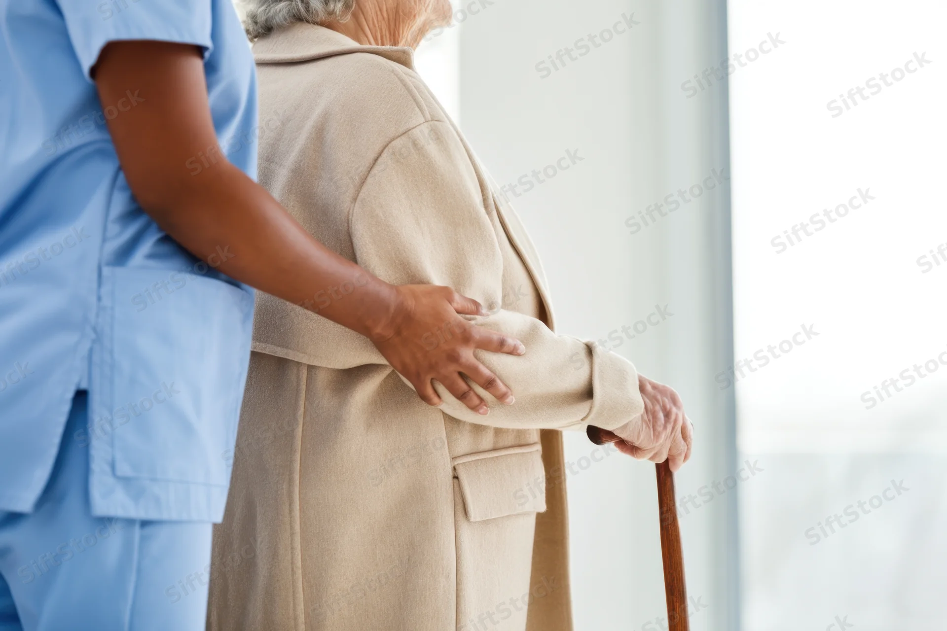 Caregiver supporting elderly person with cane while standing by window
