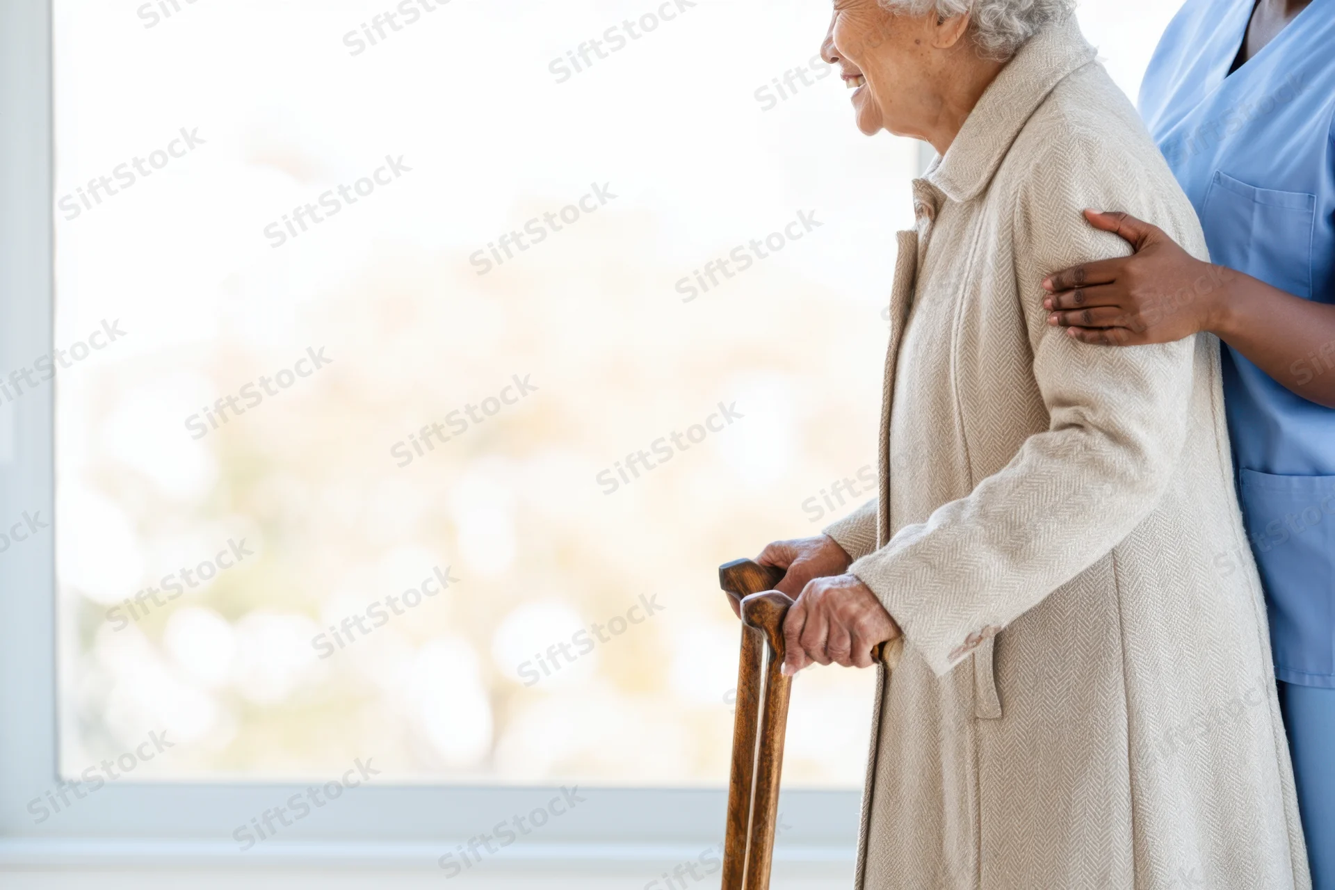Elderly woman using a cane while being supported by a caregiver near a window