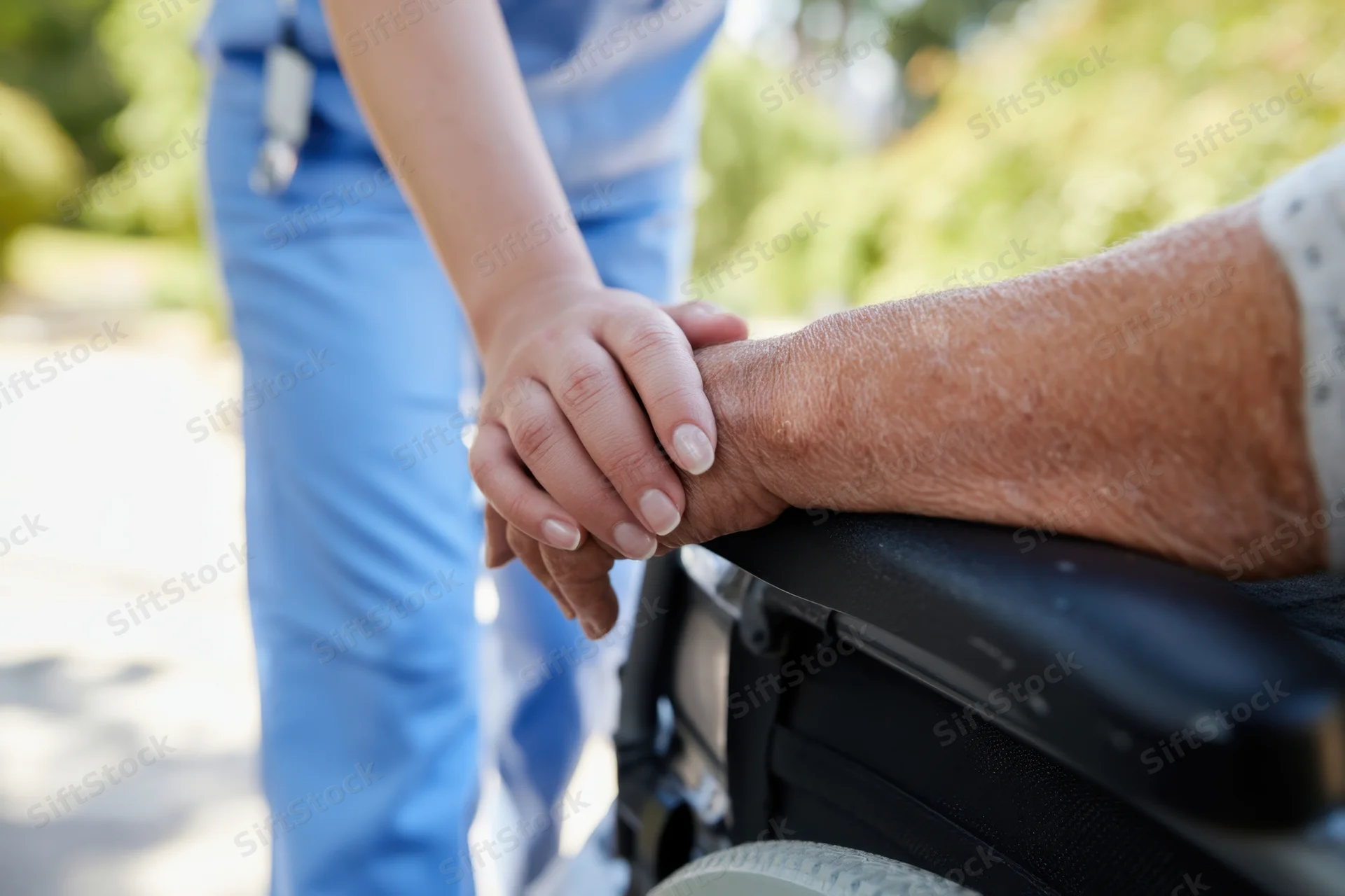 Caregiver’s hand gently holding elderly person’s hand on wheelchair armrest outdoors