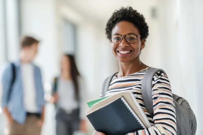 Smiling african woman student holding notebooks and wearing backpack in hallway