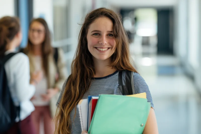 Young woman smiling while holding notebooks and folders in a school hallway