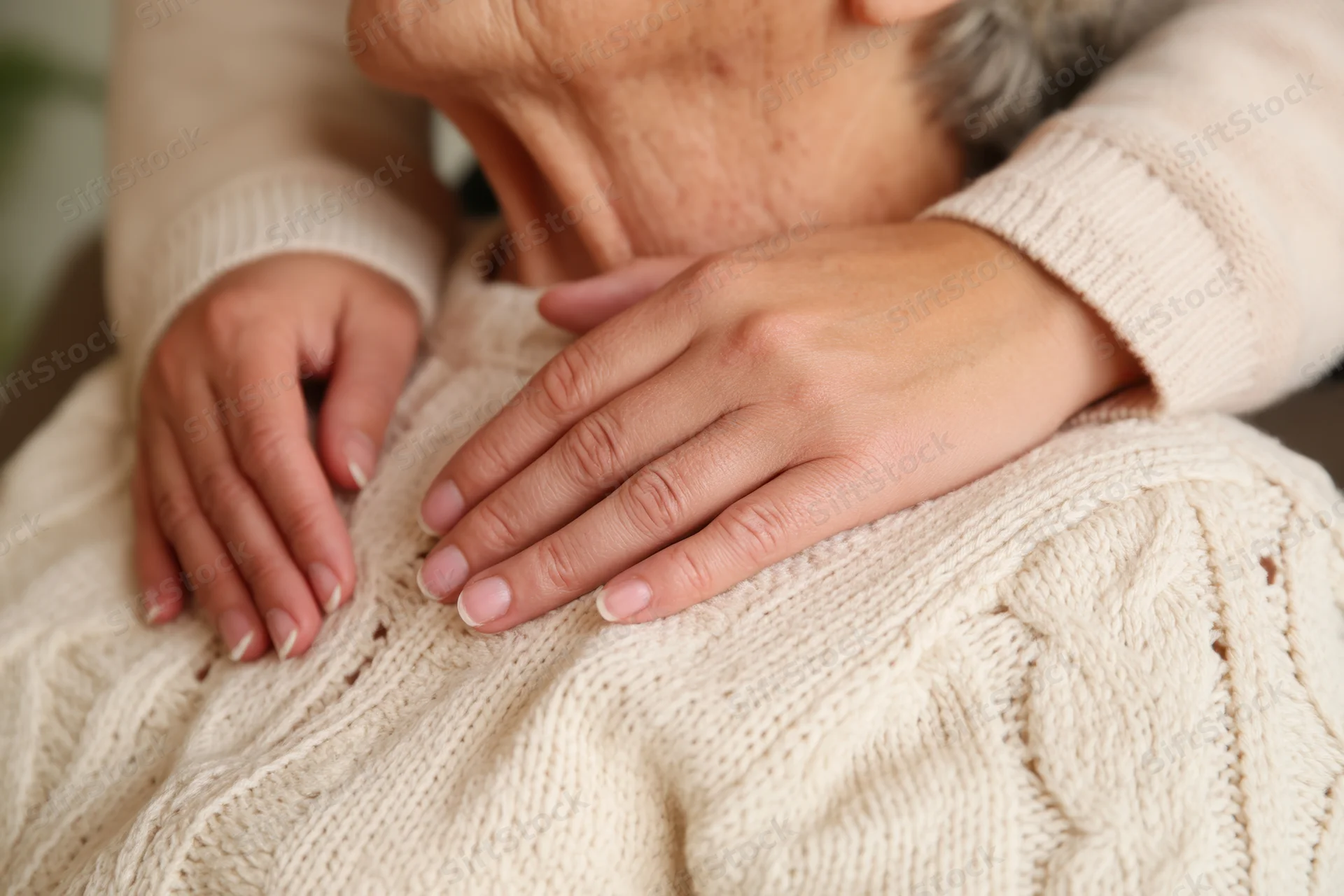 Close-up of gentle hands resting on an elderly woman's chest, wearing a cream knitted sweater